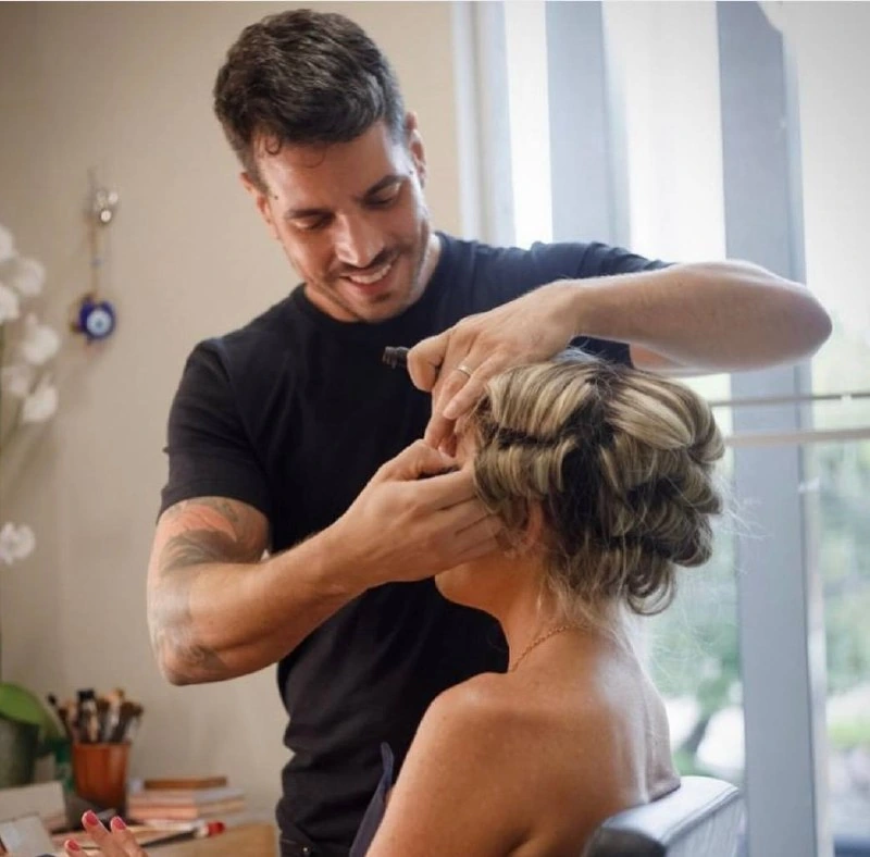 Homem em espaço de beleza ajusta o cabelo de uma mulher sentada, com itens de maquiagem ao fundo