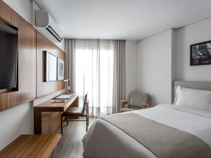 Apartamento do Nomaa Hotel com cama, escrivaninha de madeira e janela ampla (imagem ilustrativa)