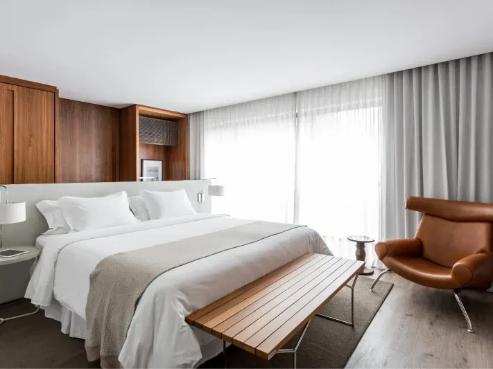Apartamento do Nomaa Hotel com cama de casal, poltrona de couro e cortinas claras (imagem ilustrativa)